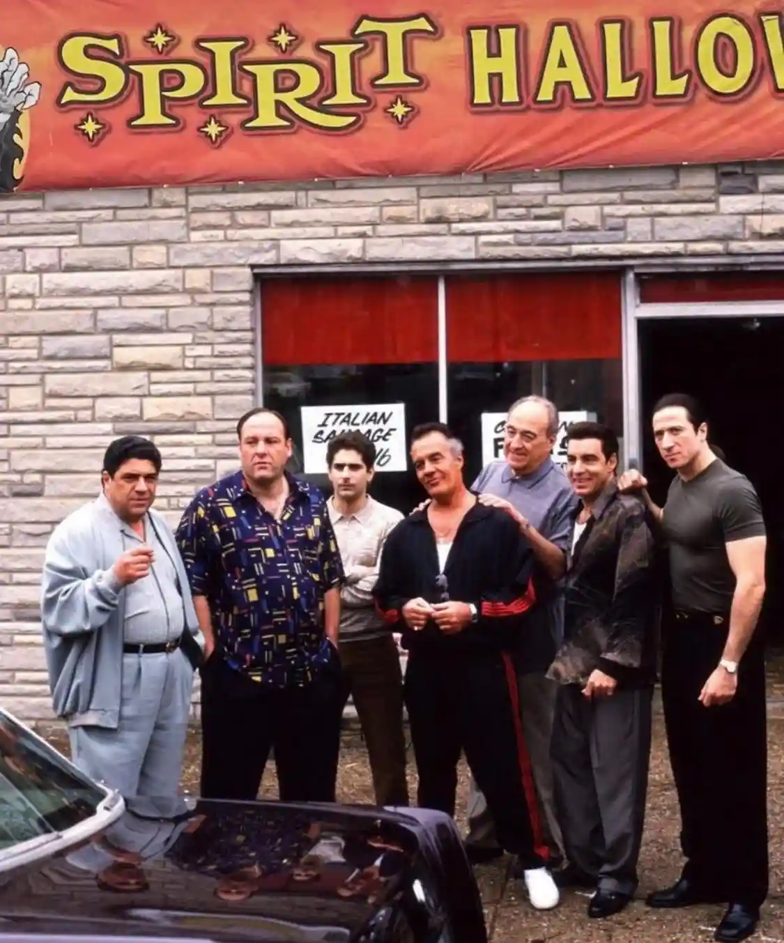 A group of seven men, recognizable as the main cast of 'The Sopranos', stand in front of a storefront with a red banner that…