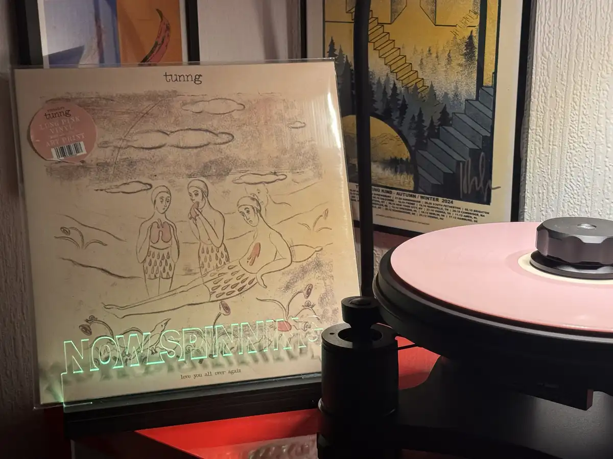 A close-up photo shows a pink vinyl record spinning on a turntable, with the album cover for 'tunng' leaning against it. The…