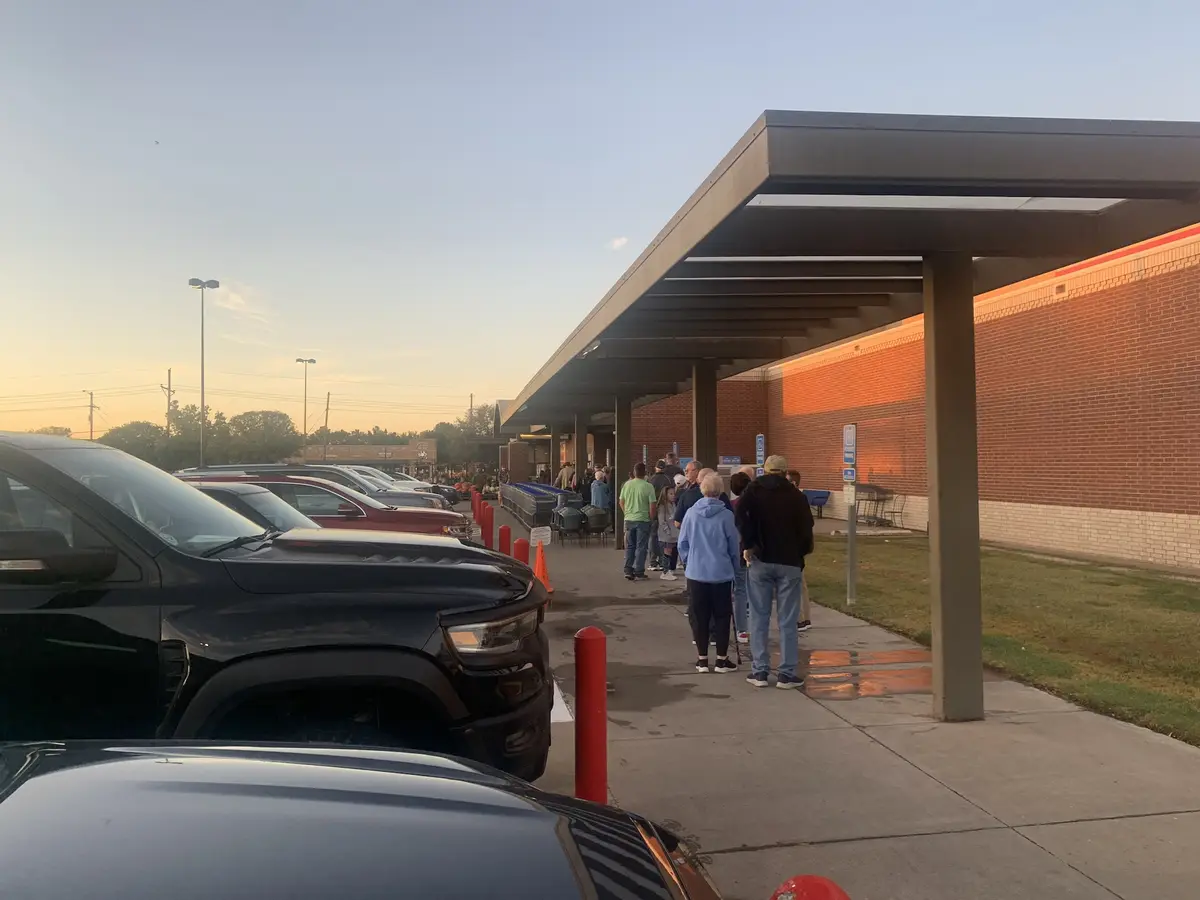 A long line of people waits outside a brick building under a covered walkway at sunset, with parked cars visible in the…