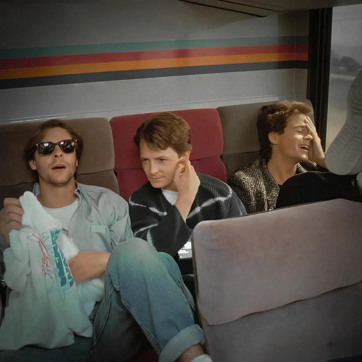 Three men sit in a bus with colorful stripes on the wall. The man on the left wears sunglasses and holds a shirt, looking…