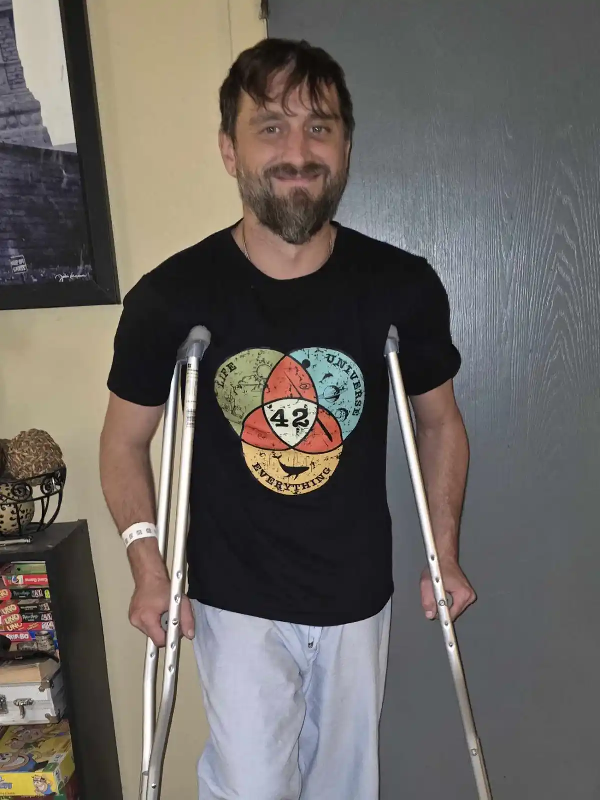 A man with a beard and crutches stands smiling, wearing a black t-shirt with a graphic that reads 'Life Universe and…