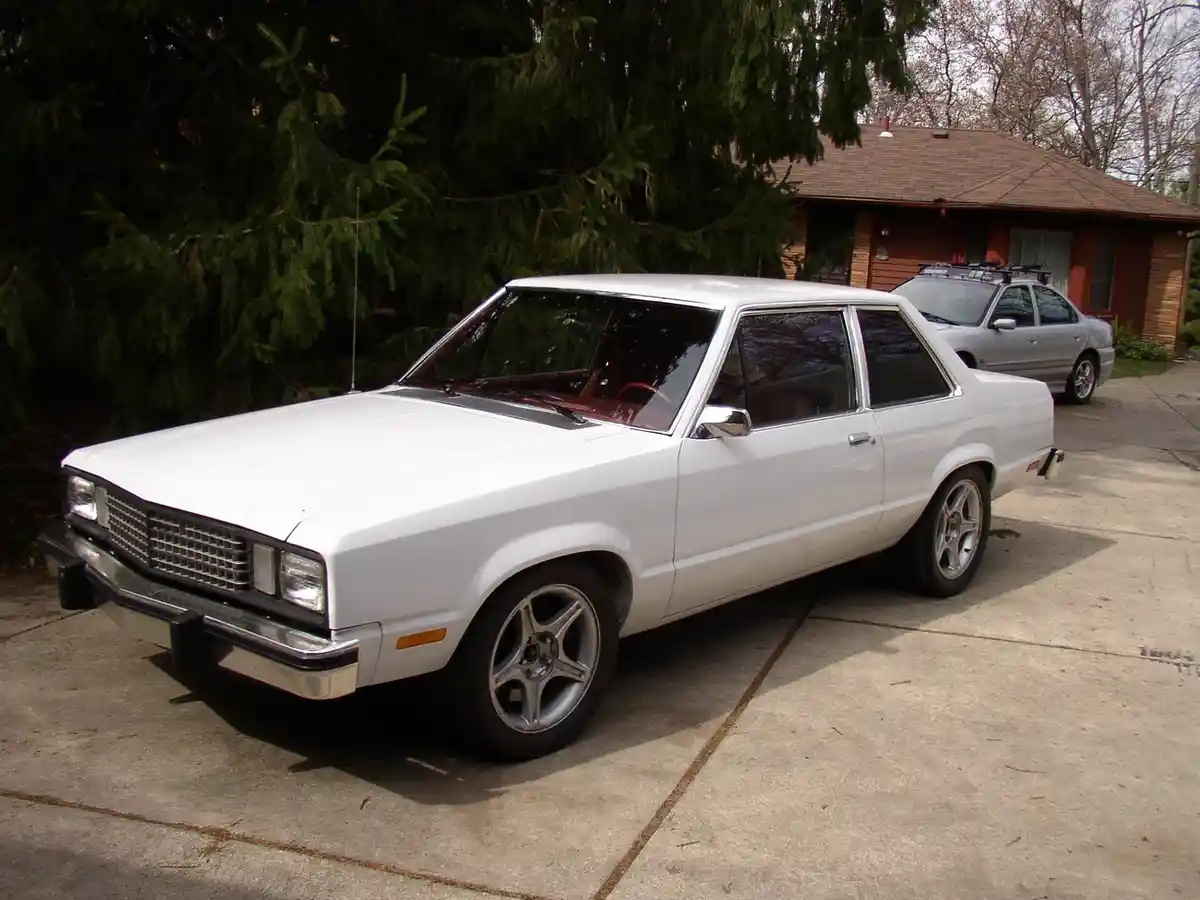 A white 1980s Chevrolet El Camino is parked in a driveway, featuring modern aftermarket silver alloy wheels and a red…