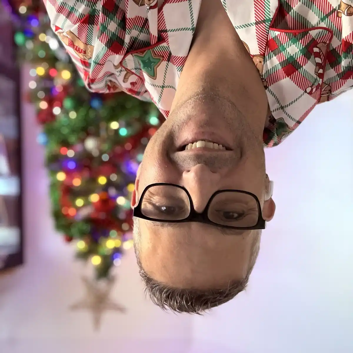 A man wearing festive red, green, and white plaid pajamas with gingerbread man and candy cane patterns is photographed…