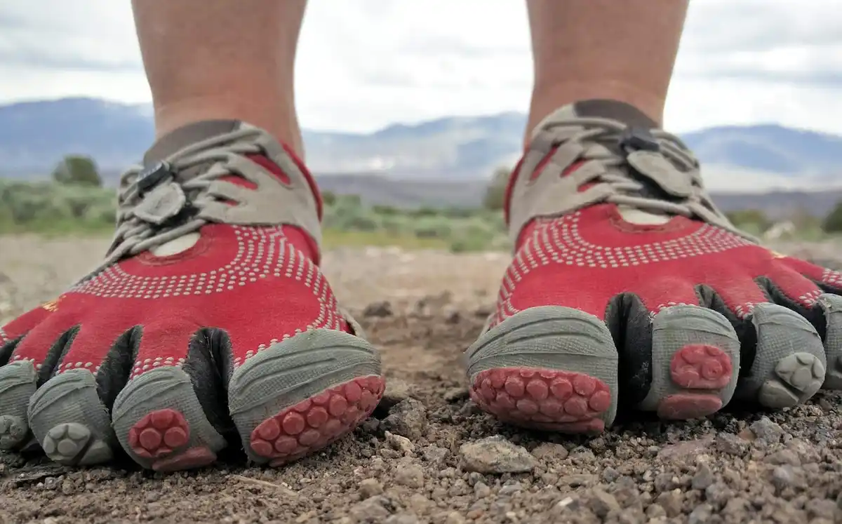 A close-up shot of a person's feet wearing red and gray toe shoes standing on gravel with mountains in the background. The…