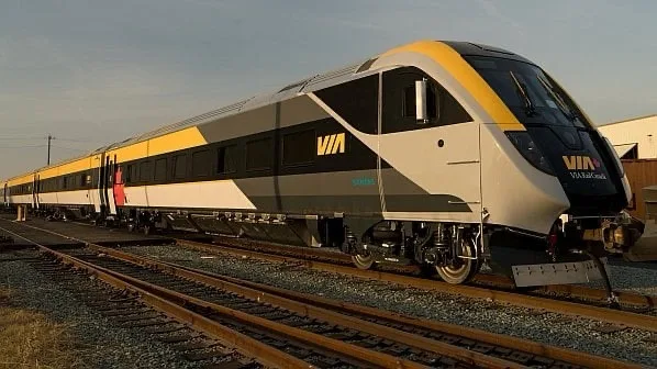 A VIA Rail passenger train with a yellow, black, and silver livery is shown on railway tracks under a clear sky. The text…