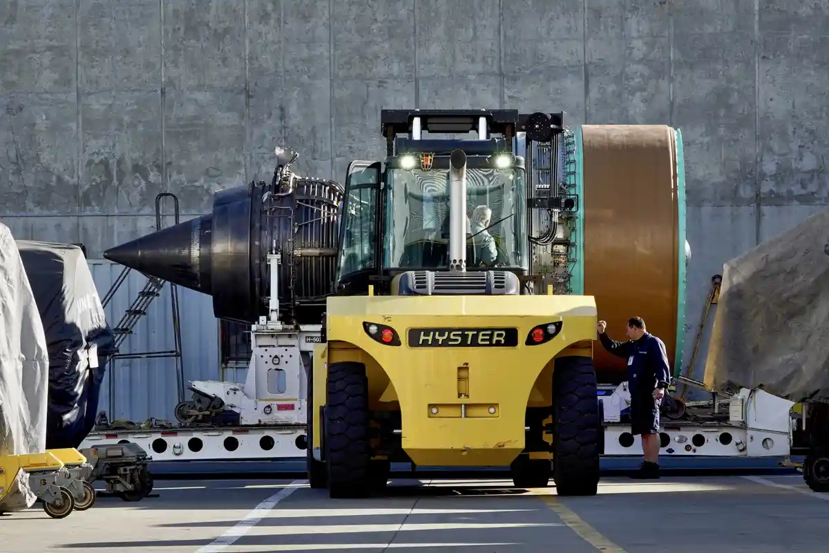A yellow Hyster forklift is shown carrying a large jet engine, with a man standing beside it and gesturing. The overlaid…
