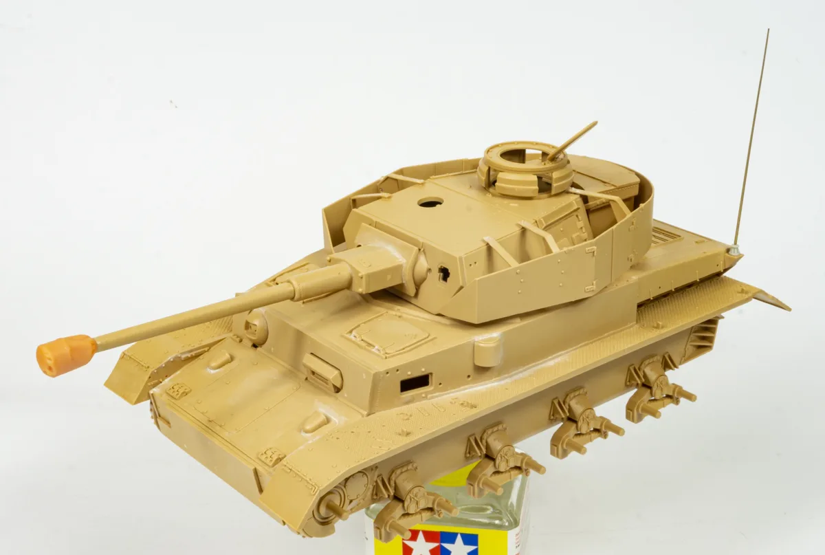 A tan plastic model tank with visible seams and unpainted details sits atop a bottle of Tamiya paint. The tank has a long…
