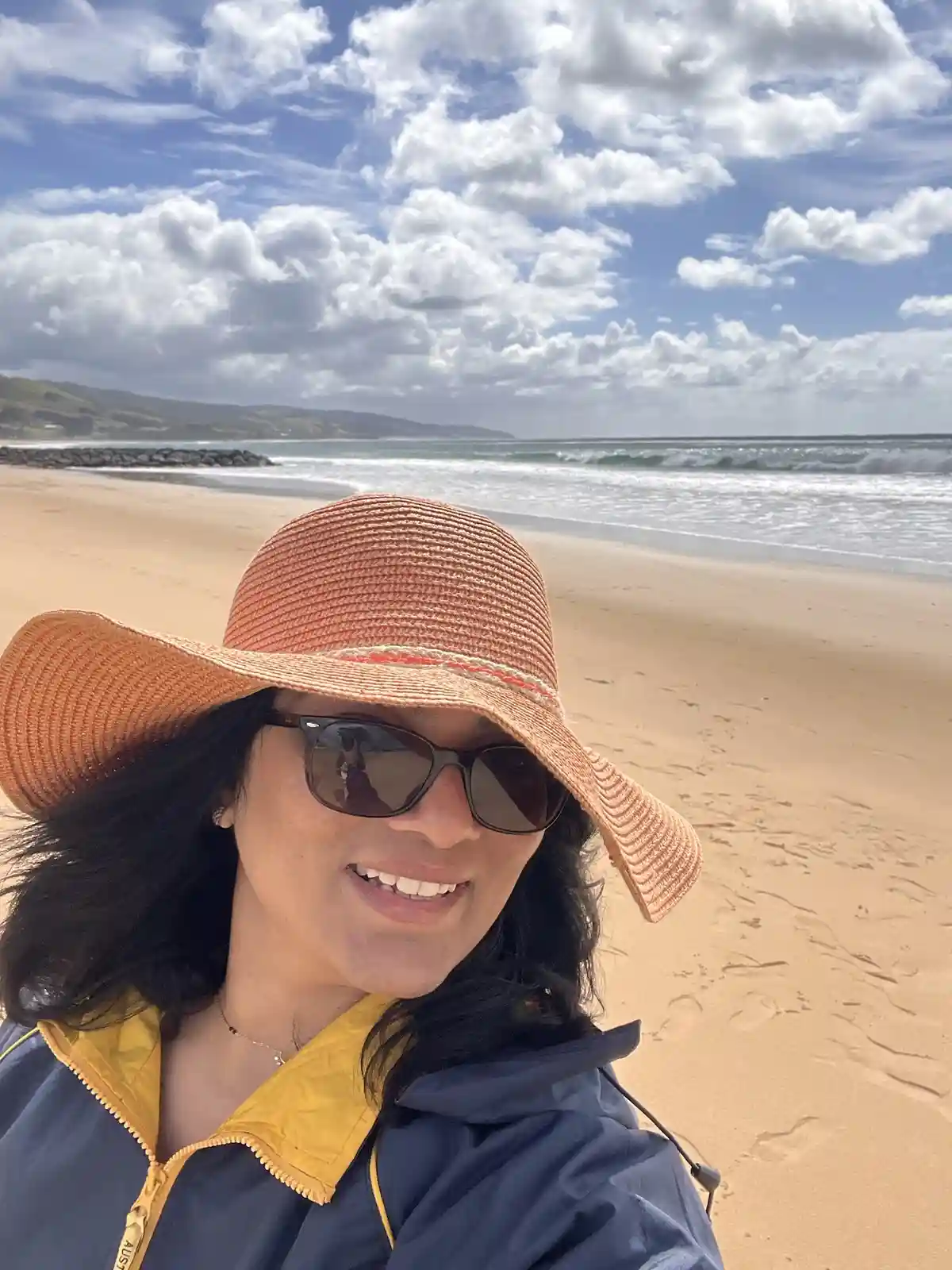 A woman with dark hair, wearing a wide-brimmed orange straw hat and dark sunglasses, smiles at the camera while taking a…