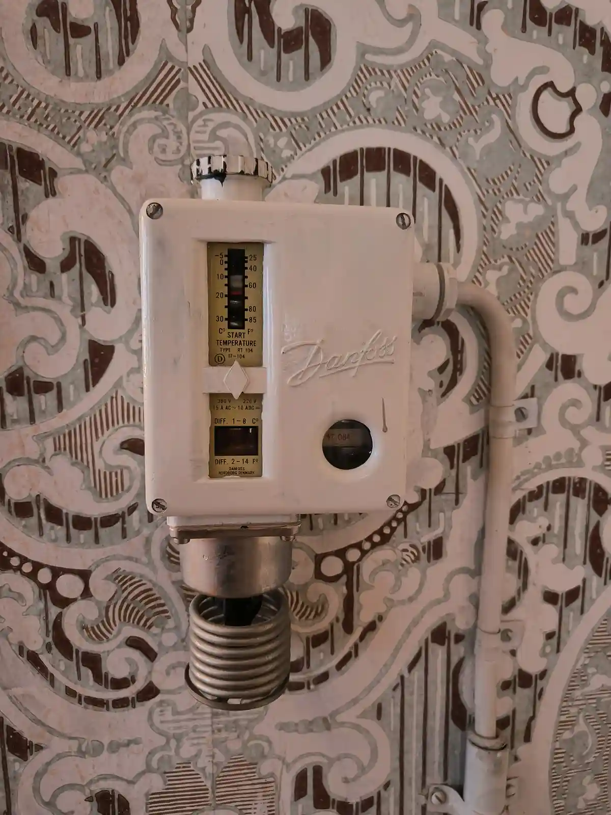 A vintage Danfoss thermostat with a temperature dial set to 60 degrees Celsius is mounted on a wall covered in ornate…