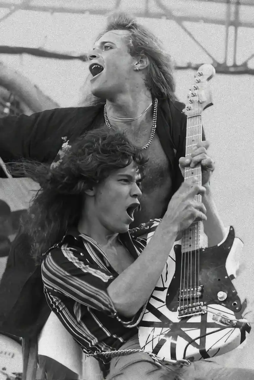 A black-and-white photo shows two male rock musicians performing energetically on stage. The frontman, with long curly hair…