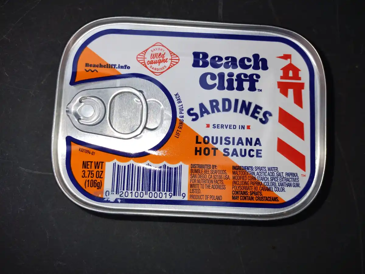 A close-up of a rectangular metal can of Beach Cliff sardines, featuring a pull-tab lid and a label with blue, white, and…