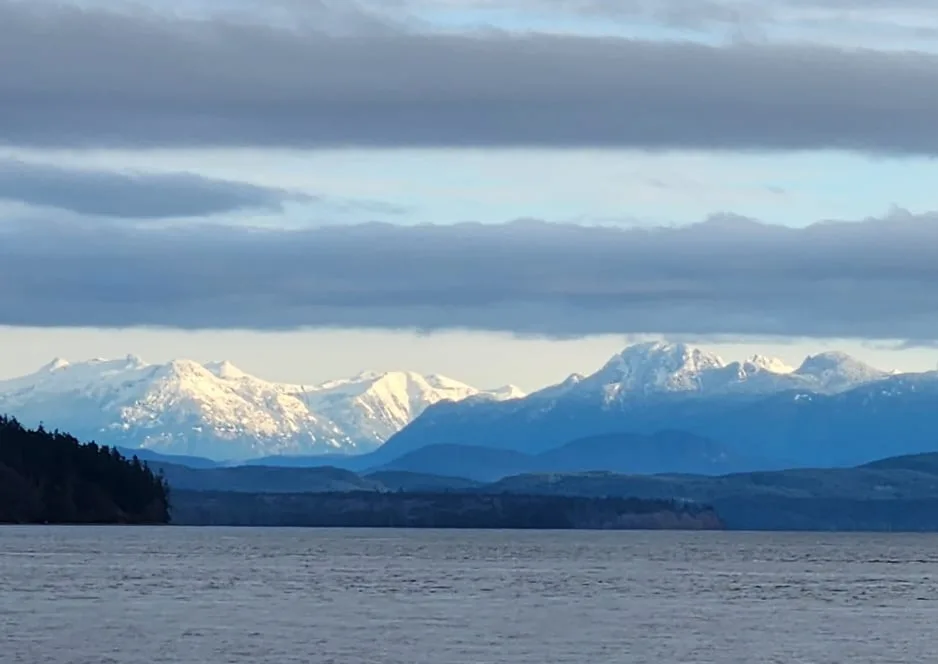 A scenic photograph shows a calm body of water in the foreground with a distant range of snow-capped mountains under a…
