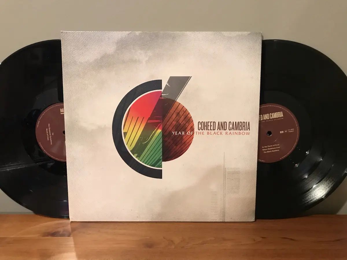 A vinyl record album by Coheed and Cambria titled 'Year of the Black Rainbow' is shown standing upright on a wooden surface…