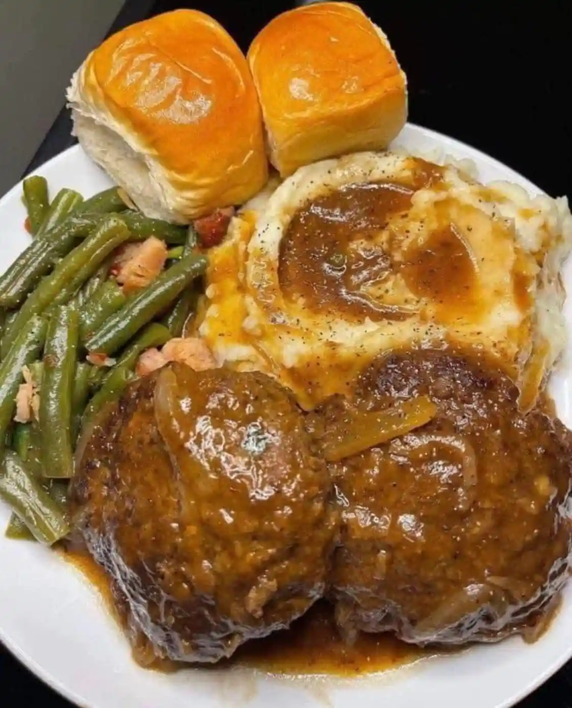A plate of home-style comfort food featuring two meatloaf patties covered in gravy, a serving of mashed potatoes with gravy…