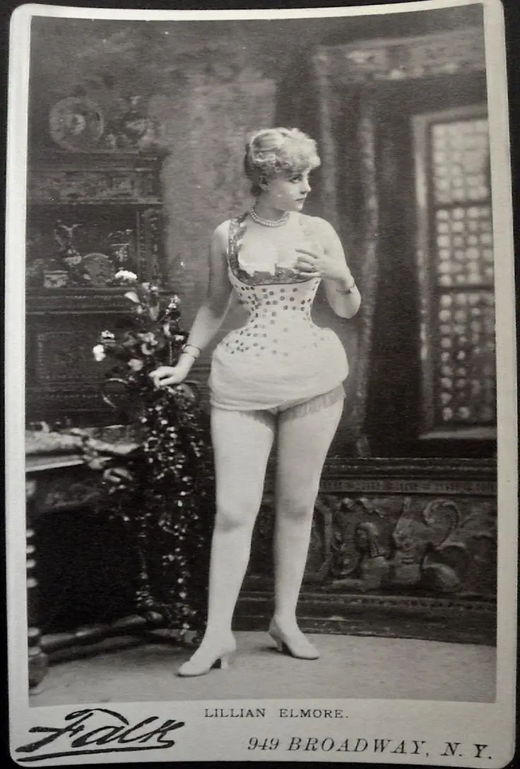 A vintage black-and-white studio portrait of a woman named Lillian Elmore, standing in an ornate room, wearing a revealing…