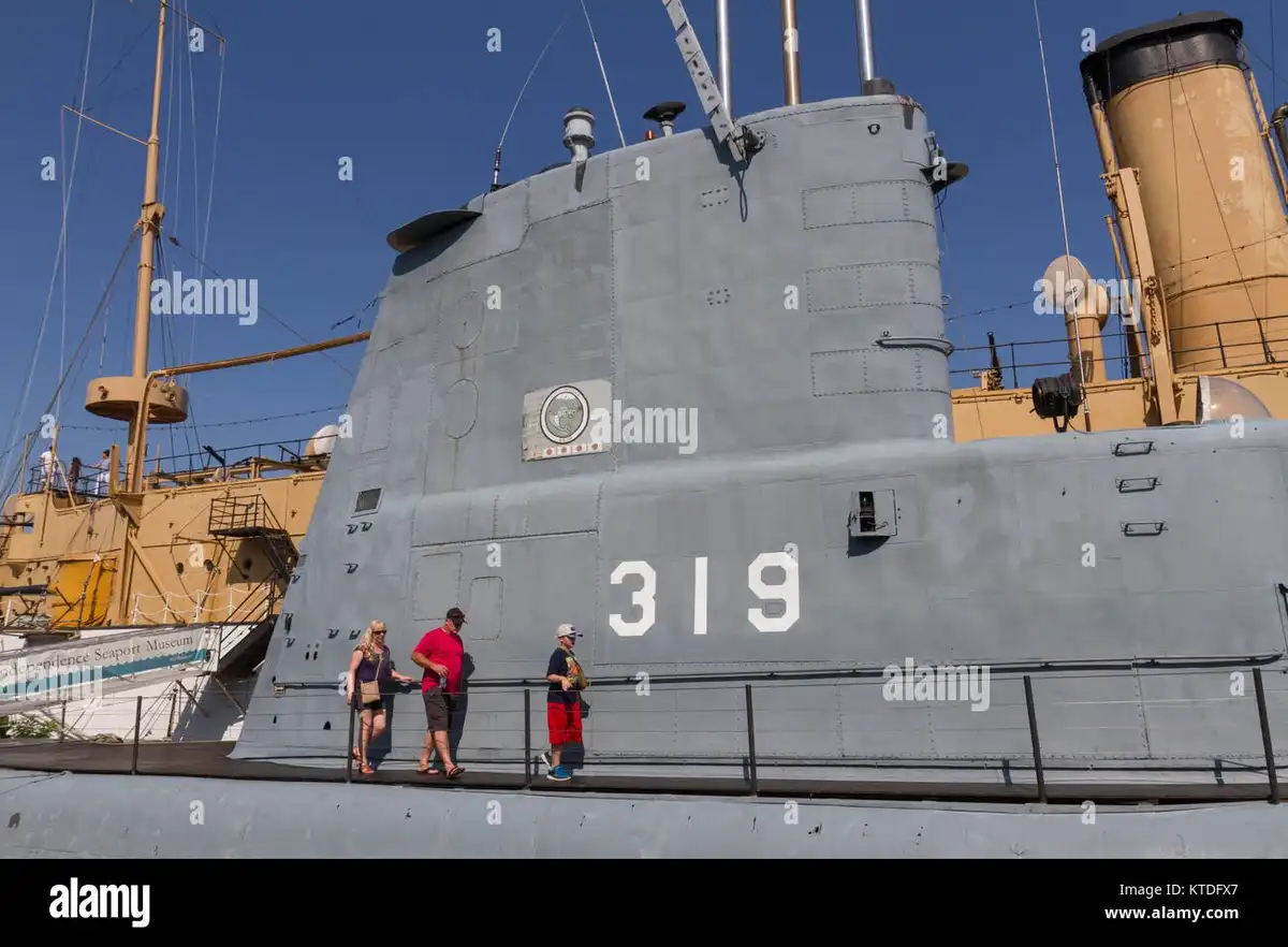 A gray submarine with the number 319 painted in large white numerals on its side is shown under a clear blue sky. Three…