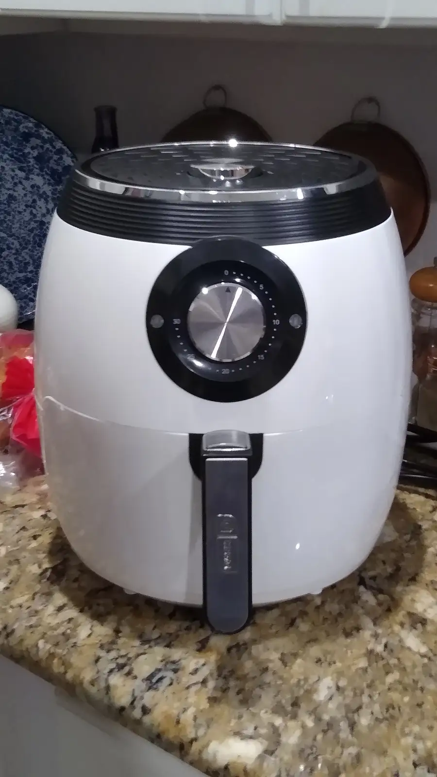 A white air fryer with a black dial and handle sits on a granite countertop in a kitchen. The dial is set to zero, and the…