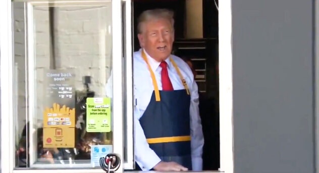 A man with gray hair wearing a white shirt, red tie, and a blue and yellow apron peers out from a window of a fast-food…