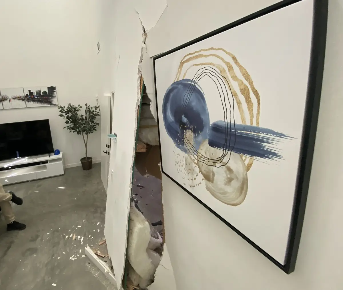 A large abstract painting with blue and gold swirls hangs on a wall that has been partially demolished, revealing a messy…
