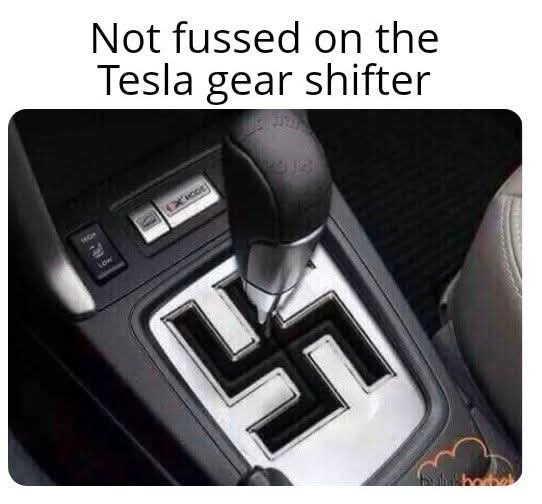 A close-up photo of a Tesla car's center console shows a gear shifter with a gear pattern that is shaped like a swastika…