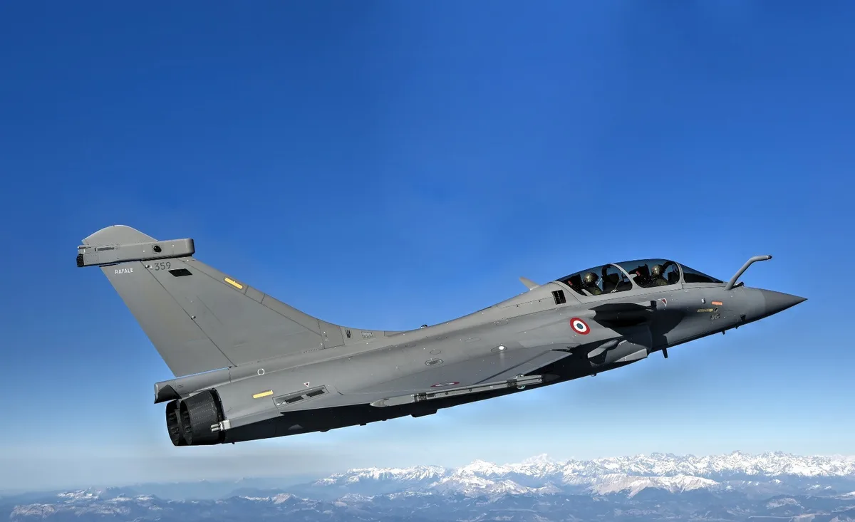 A grey French Dassault Rafale fighter jet is shown in flight against a clear blue sky, with snow-capped mountains visible…
