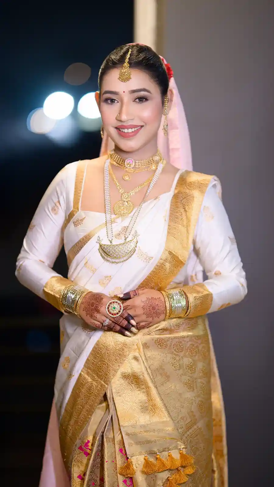 A woman in traditional Indian bridal attire, wearing a white and gold saree, heavy gold jewelry, and henna on her hands…