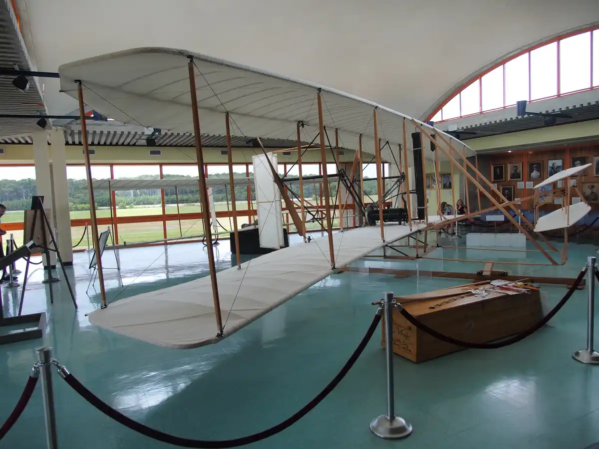 a large, early 20th-century biplane with wooden frame and white fabric wings is displayed indoors on a polished teal floor…