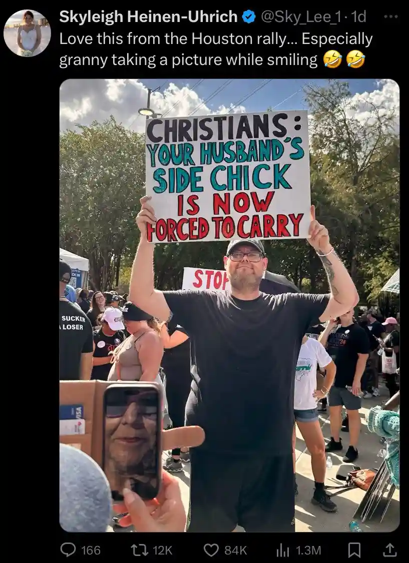 A man at an outdoor rally holds up a handmade protest sign that reads 'CHRISTIANS: YOUR HUSBAND'S SIDE CHICK IS NOW FORCED…