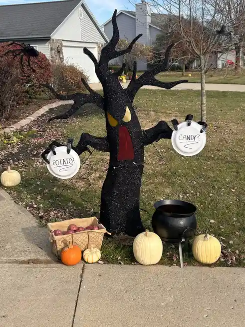 A black, glittery Halloween witch tree decoration with yellow eyes and a red mouth stands in a front yard, holding two white…
