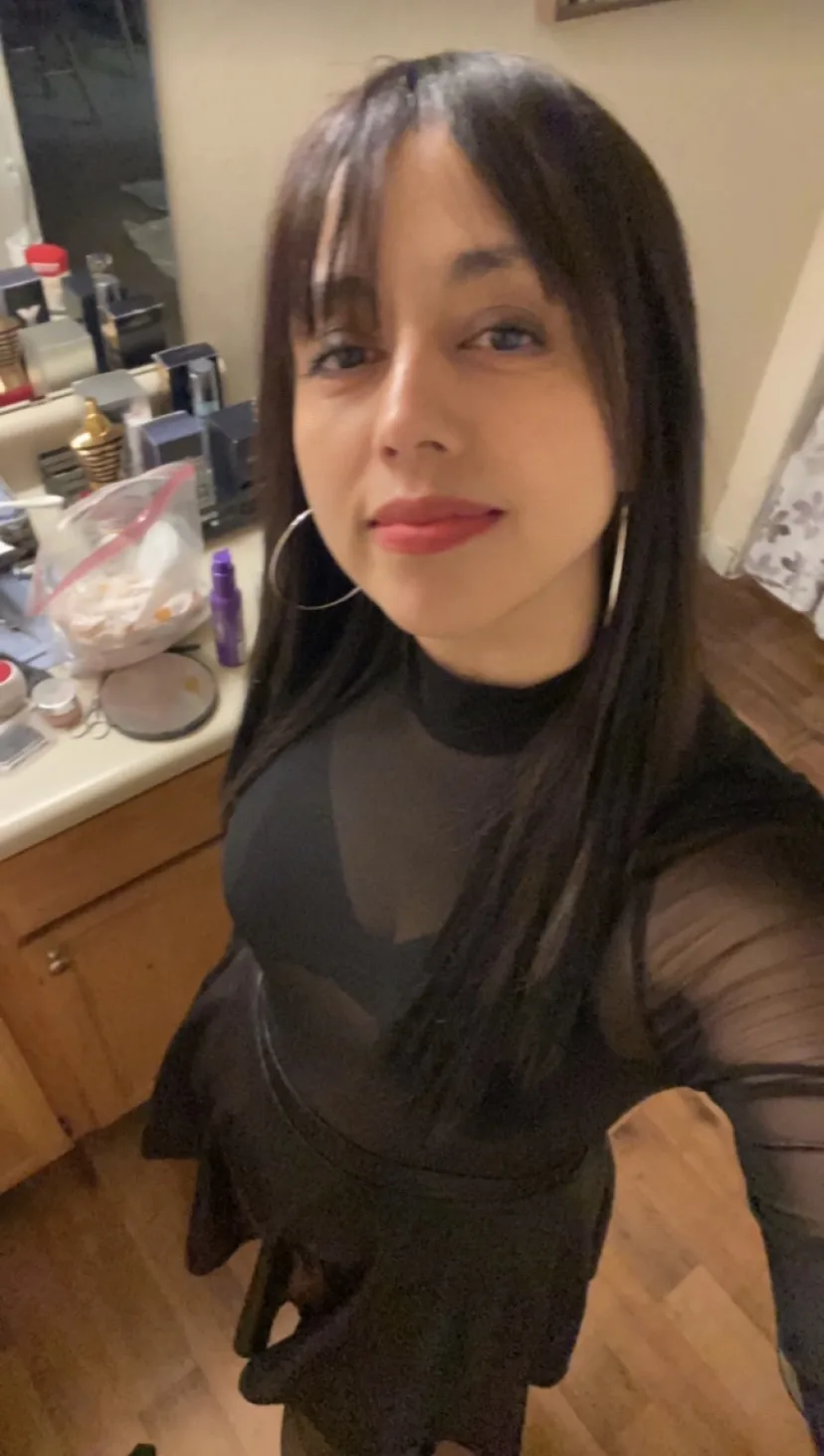 A selfie of a woman with long dark hair and bangs, wearing a black sheer top and large hoop earrings, standing in a room…