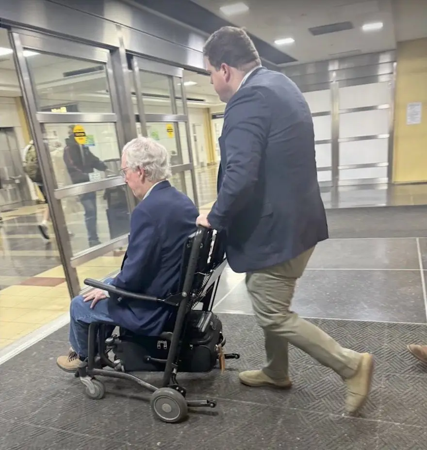 A man in a dark suit pushes an older gentleman in a motorized wheelchair through a modern building with glass doors. The man…