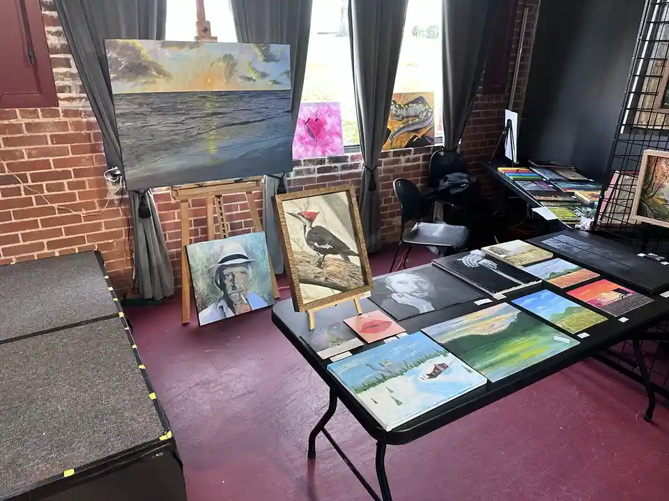 A room with a red brick wall and windows draped with grey curtains, displaying numerous paintings on easels and tables. The…