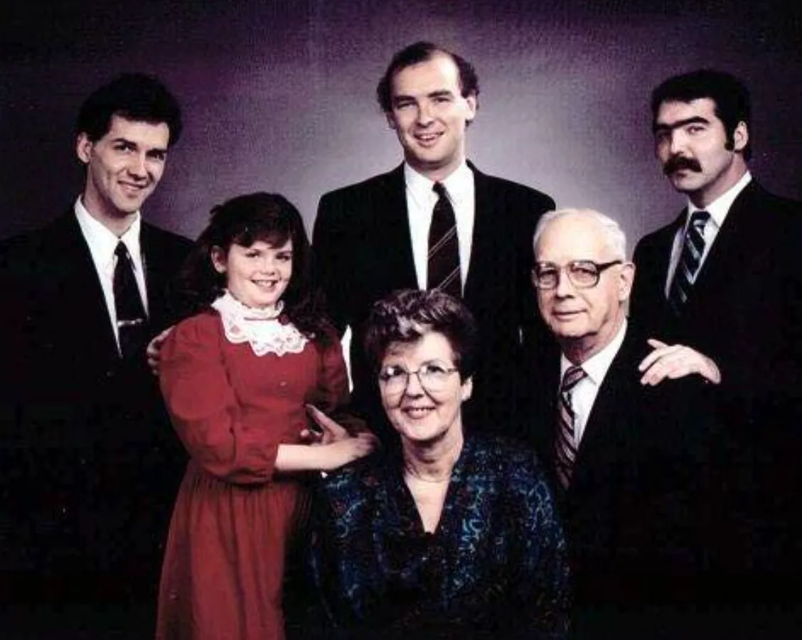 A vintage family portrait shows six people posing formally: a young girl in a red dress, an older woman in a dark patterned…