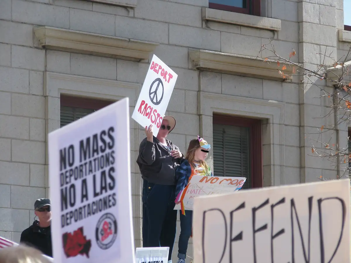 A protest scene with multiple signs, one prominently reading 'DEPORT RACISTS' with a peace symbol, another saying 'NO MASS…