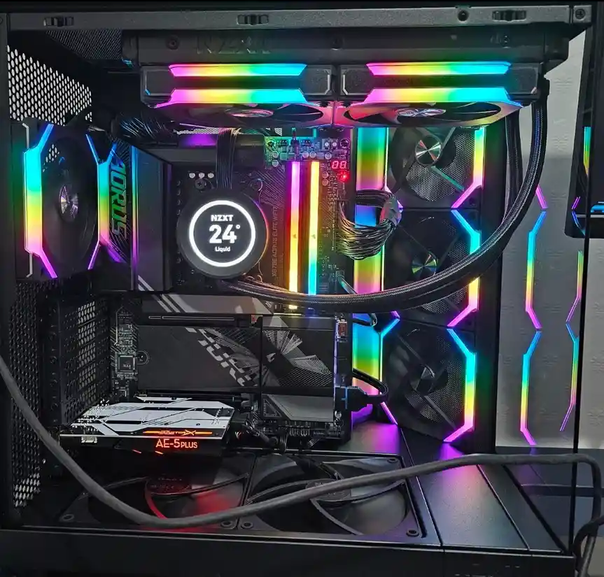 A high-end custom PC build with rainbow RGB lighting on fans and case, showing an NZXT liquid cooler displaying '24° Liquid'…