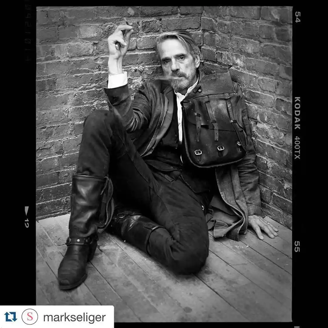 A black-and-white photo of a man with a beard and mustache, sitting on a wooden floor against a brick wall, wearing a…