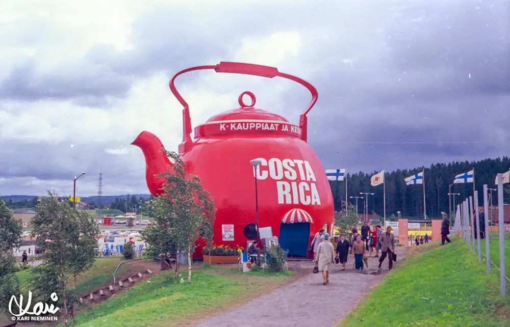 A giant red teapot structure with 'COSTA RICA' written on its side and 'K-KAUPPIAAT JA KESKUSTA' on its lid, set outdoors…