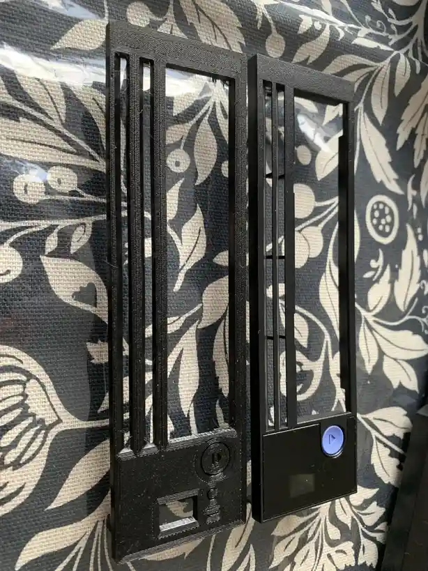Two black 3D-printed objects resembling old iPod Classics sit side by side against a black-and-white floral patterned…