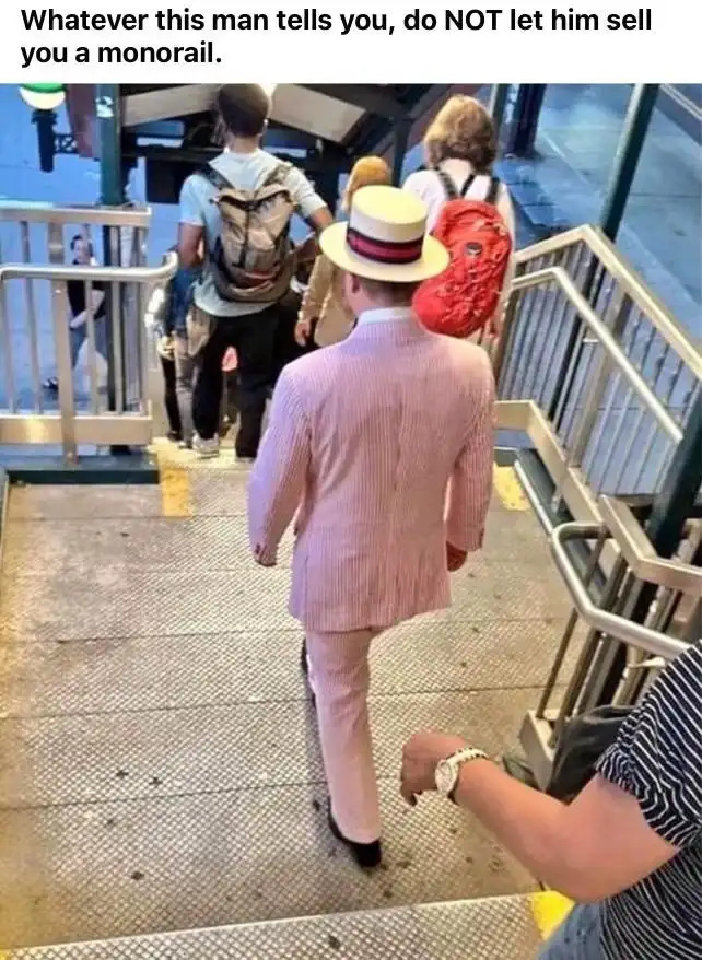A man in a pink pinstripe suit and a white hat with a red band walks down a metal staircase in a transit station, viewed…