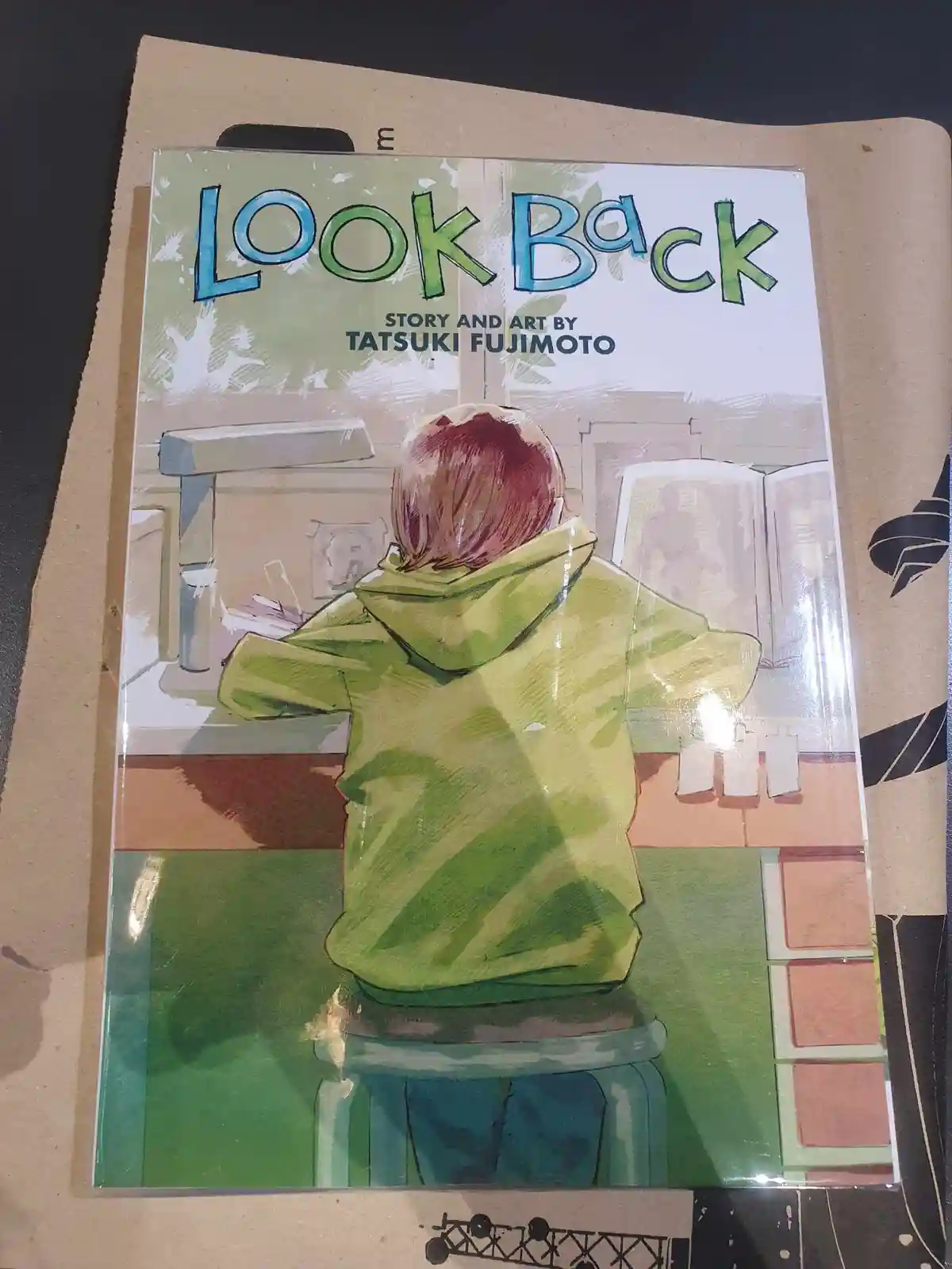 The image shows the cover of the manga 'Look Back' by Tatsuki Fujimoto, depicting a person with short red hair in a green…