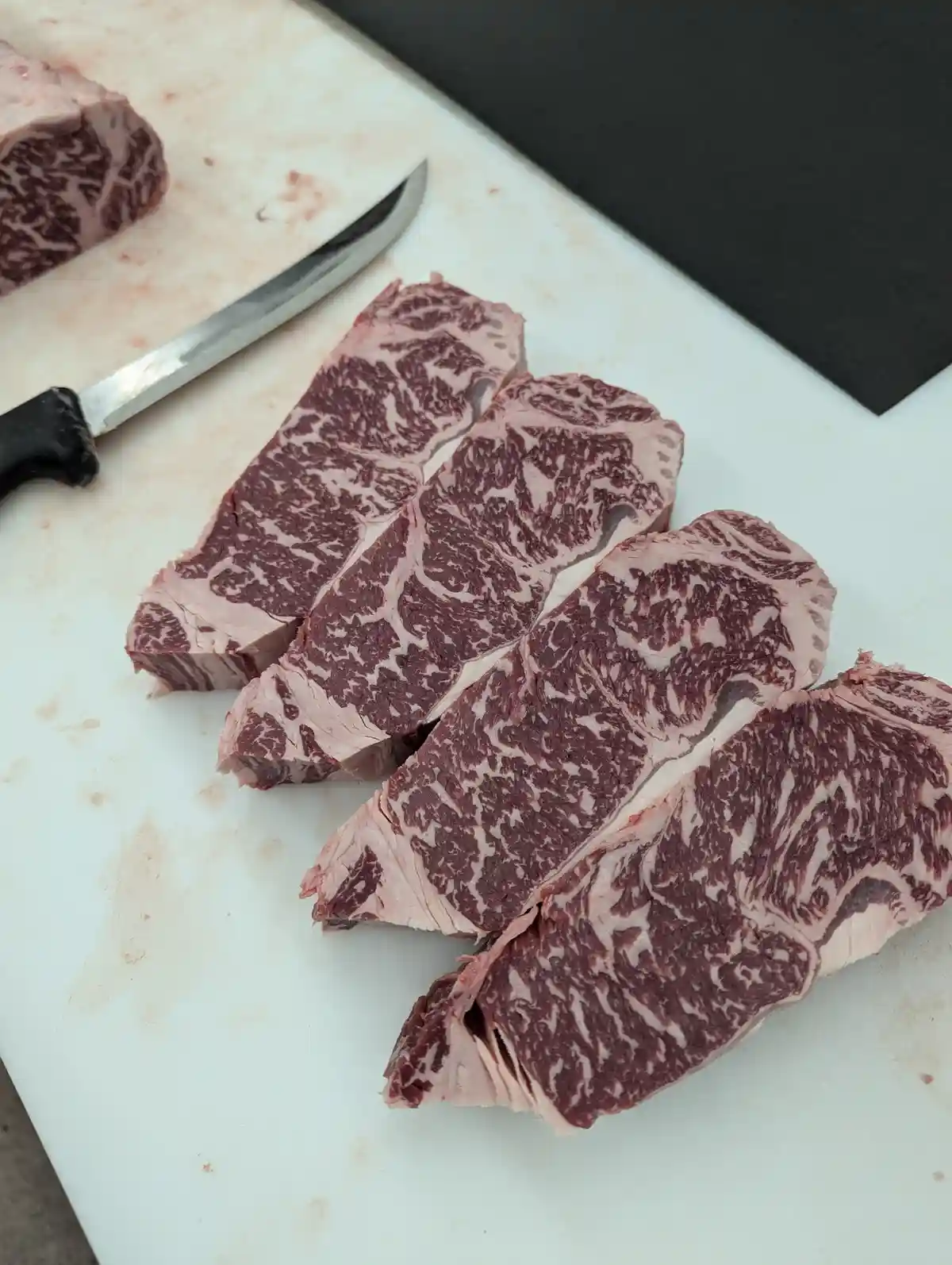 A close-up photo shows several thick slices of high-grade marbled beef arranged on a white cutting board, with a knife…