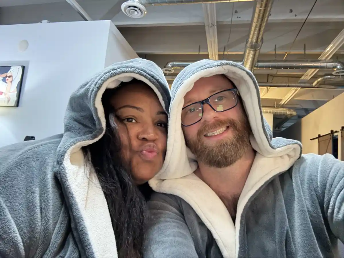 A selfie of a man and a woman both wearing identical grey fleece hooded robes with white lining, taking a selfie in what…