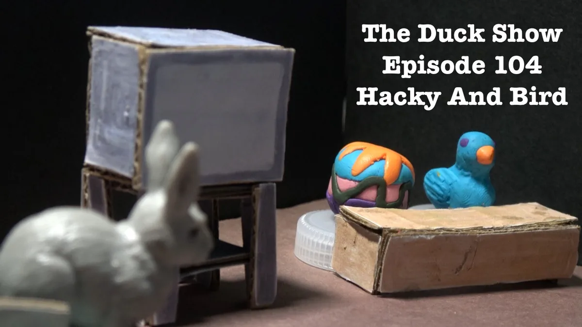 A stop-motion style diorama features a white rabbit figurine watching a cardboard TV set, while a blue clay bird and a…