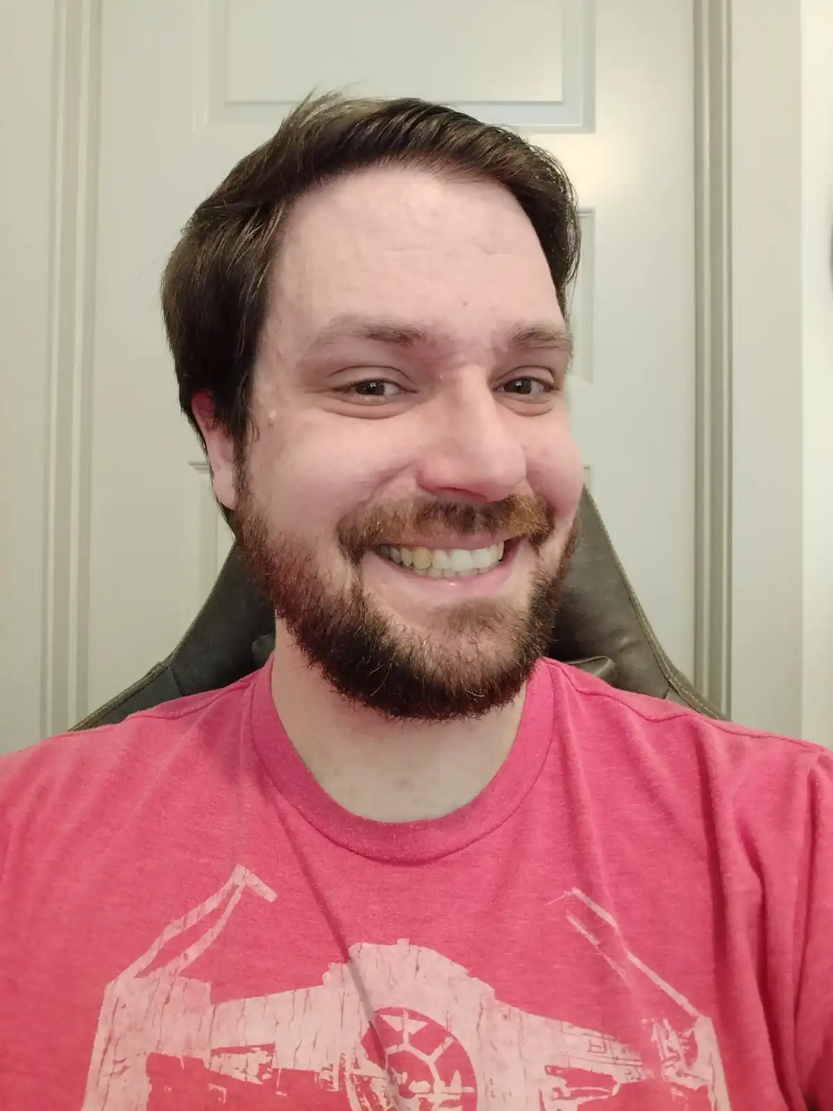 A man with a brown beard and short brown hair smiles broadly at the camera while wearing a pink t-shirt with a faded graphic…