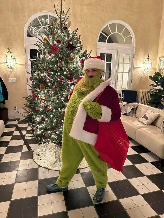 A person in a full-body green Grinch costume, complete with a Santa hat and a red velvet sack that reads 'Merry Christmas'…