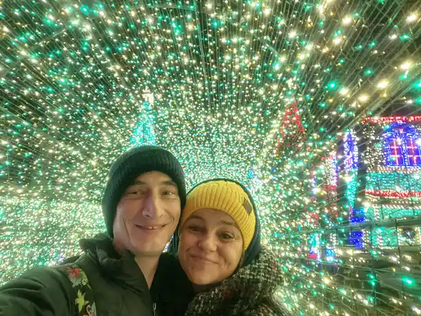 A selfie of a smiling man and woman wearing winter hats, posing in front of a dazzling canopy of green and white string…