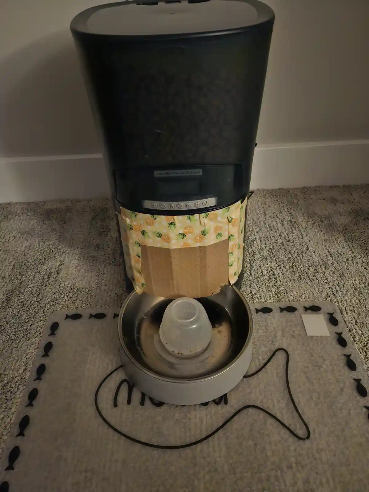 A black HoneyGuardian pet feeder stands on a carpet, with a cardboard tube and pineapple-patterned fabric wrapped around its…
