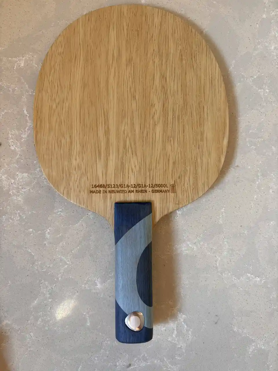 A close-up photo shows a wooden table tennis paddle resting on a light-colored speckled countertop. The paddle has a light…