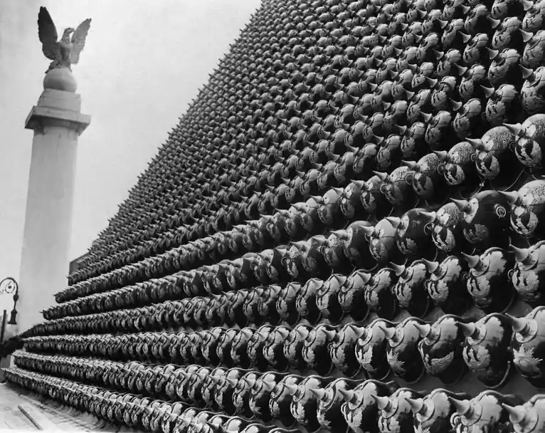 A black-and-white historical photograph shows a steeply sloped roof covered in hundreds of ornate, spiked military helmets…