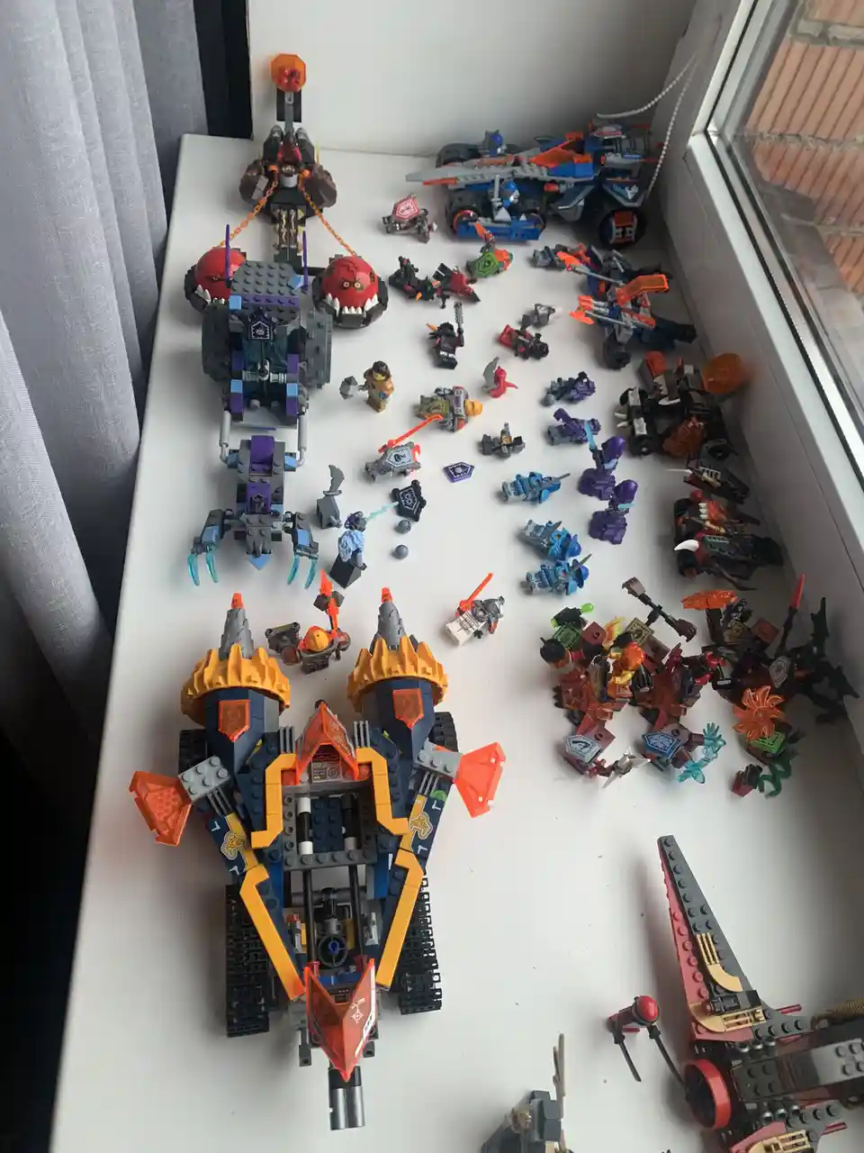 A white windowsill is covered with numerous Lego Ninjago sets and minifigures, including large vehicles and scattered…